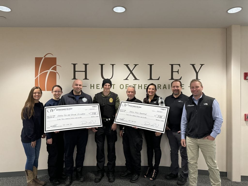 Whitaker Marketing Group presents donation checks to the Huxley Police Department and Huxley Fire and Rescue, with team members standing together at the Huxley City Hall.