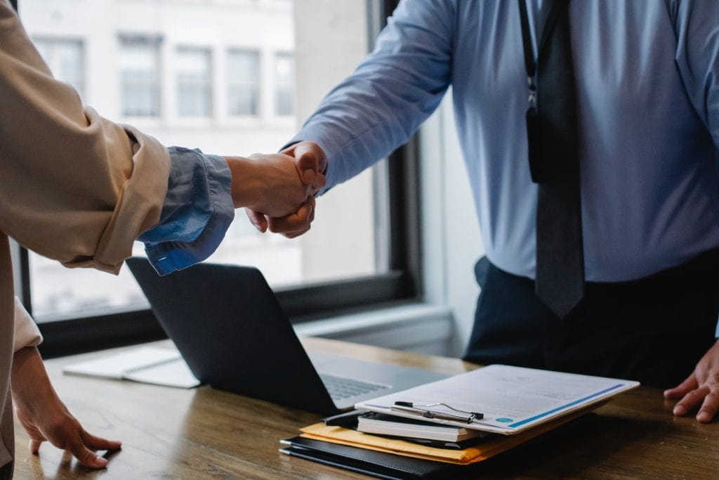 A real estate professional and a client shaking hands across a desk with documents and a laptop in the background.