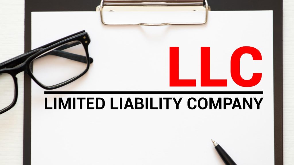 Clipboard with the words LLC Limited Liability Company, glasses, and a pen on a desk.