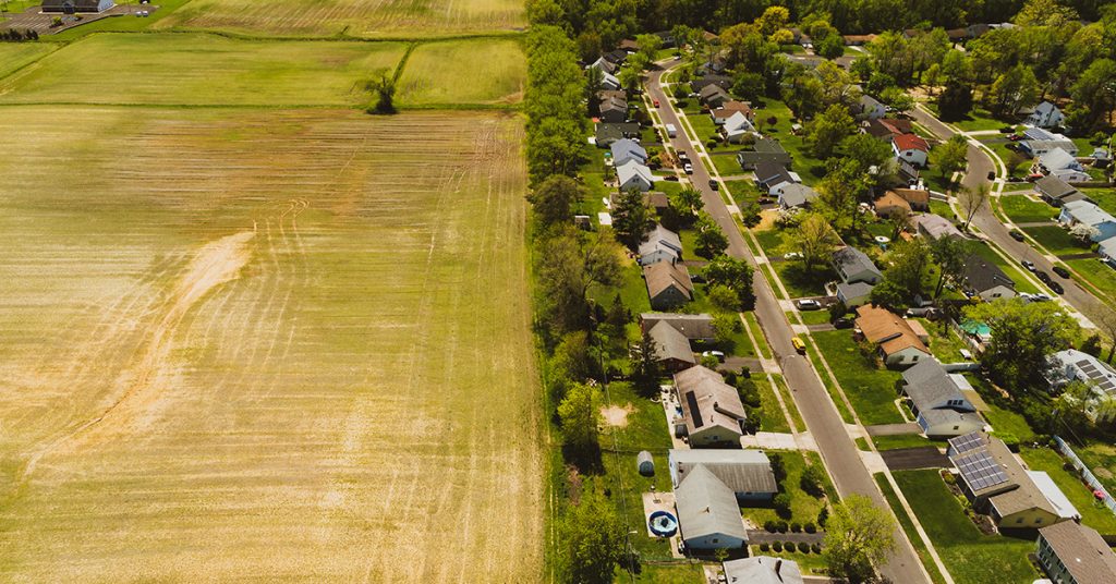 Aerial view showing a clear divide between agricultural farmland and a suburban residential neighborhood, highlighting the impact of zoning boundaries.