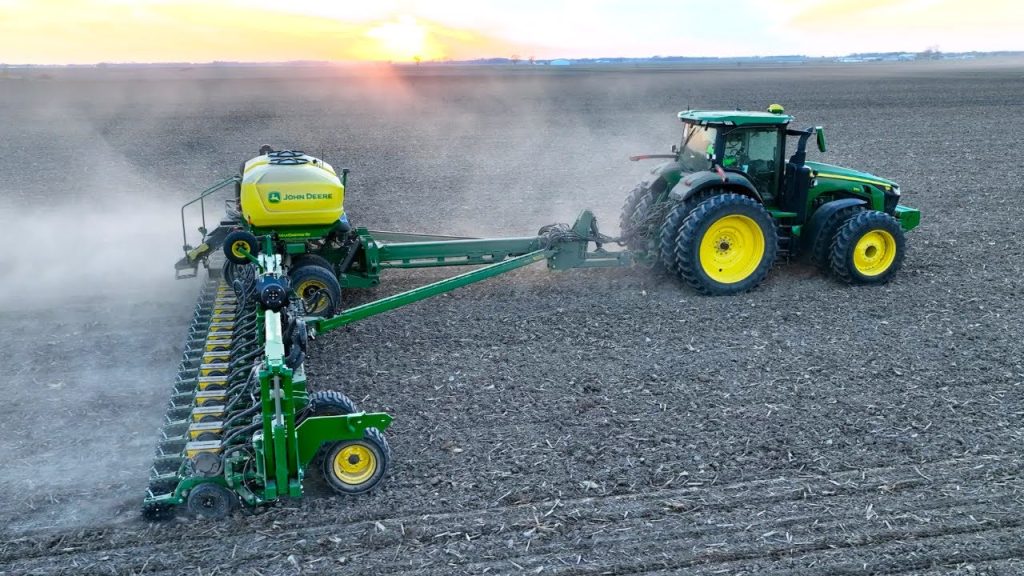 A John Deere tractor pulling a planter across a large field during spring planting at sunset, kicking up dust as it seeds the soil.