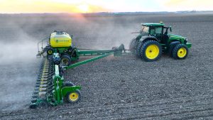A John Deere tractor pulling a planter across a large field during spring planting at sunset, kicking up dust as it seeds the soil.