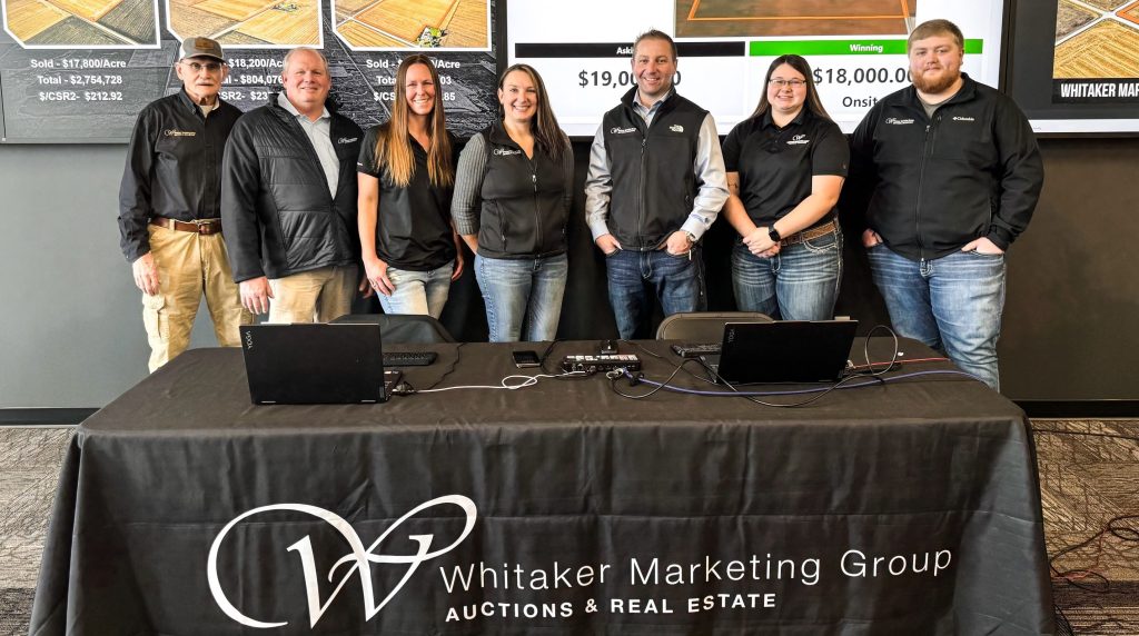 Team of auction and real estate professionals at Whitaker Marketing Group standing behind a conference table with branding.
