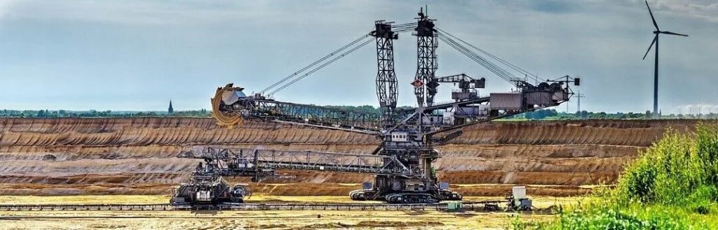 Large mining excavator operating in an open-pit extraction site with visible soil layers and a wind turbine in the background.