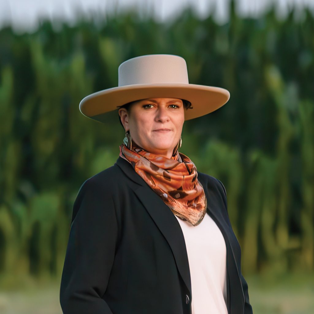 Kathy Loew standing outdoors in front of a green field, wearing a wide-brim hat and blazer