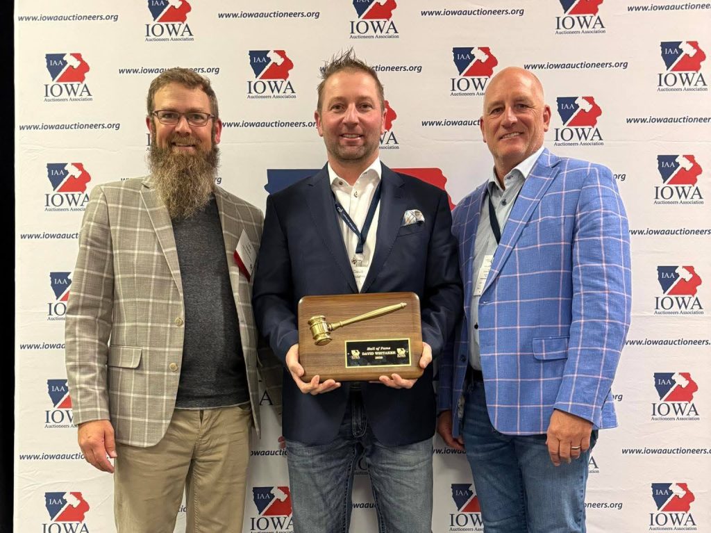 Three auctioneers standing in front of an Iowa Auctioneers Association backdrop, with the center recipient holding a wooden plaque featuring a ceremonial gavel award.